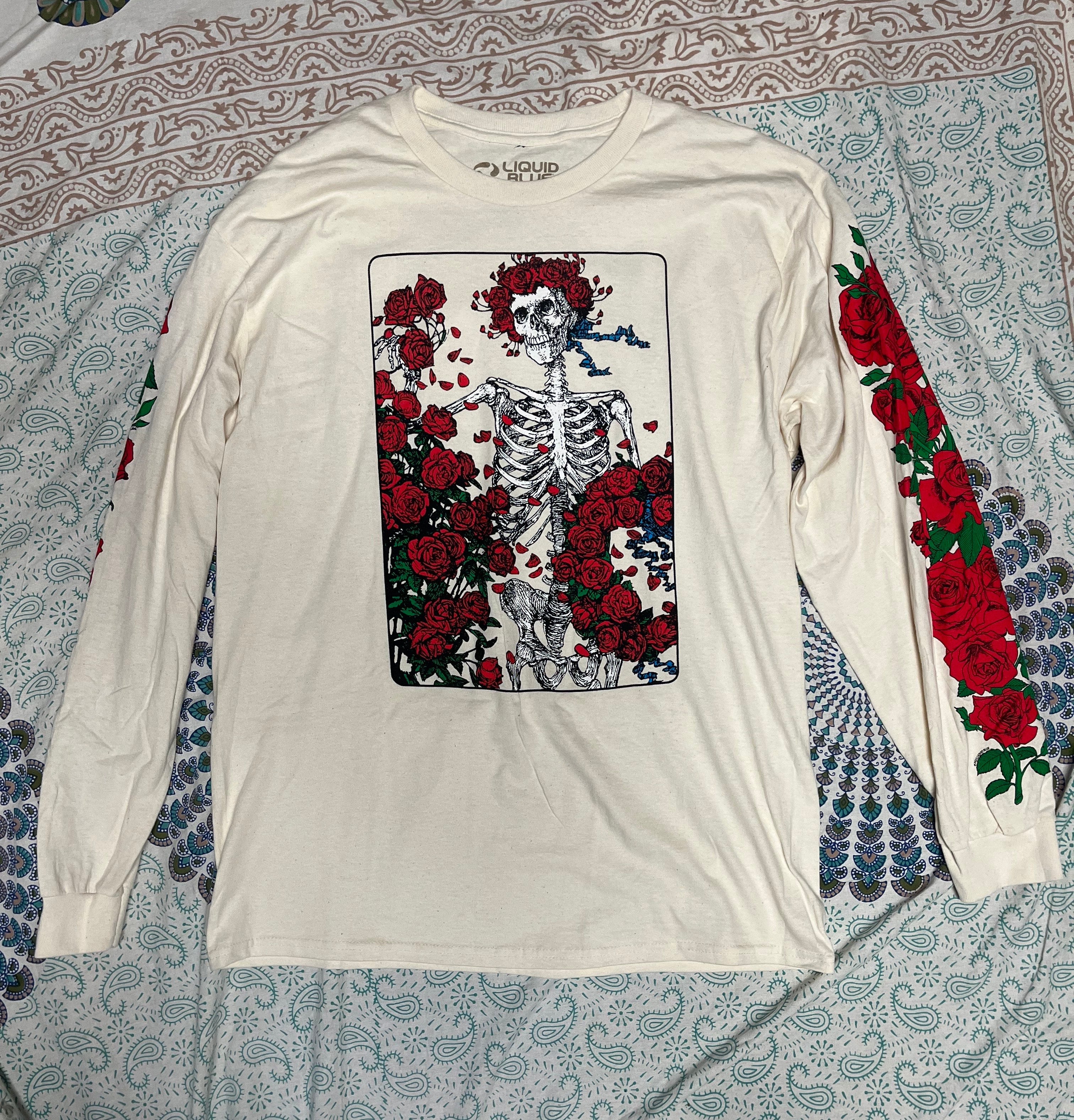 White Grateful Dead longsleeve shirt with skeleton and rose design on a patterned fabric background. Front of shirt. From company Liquid Blue.