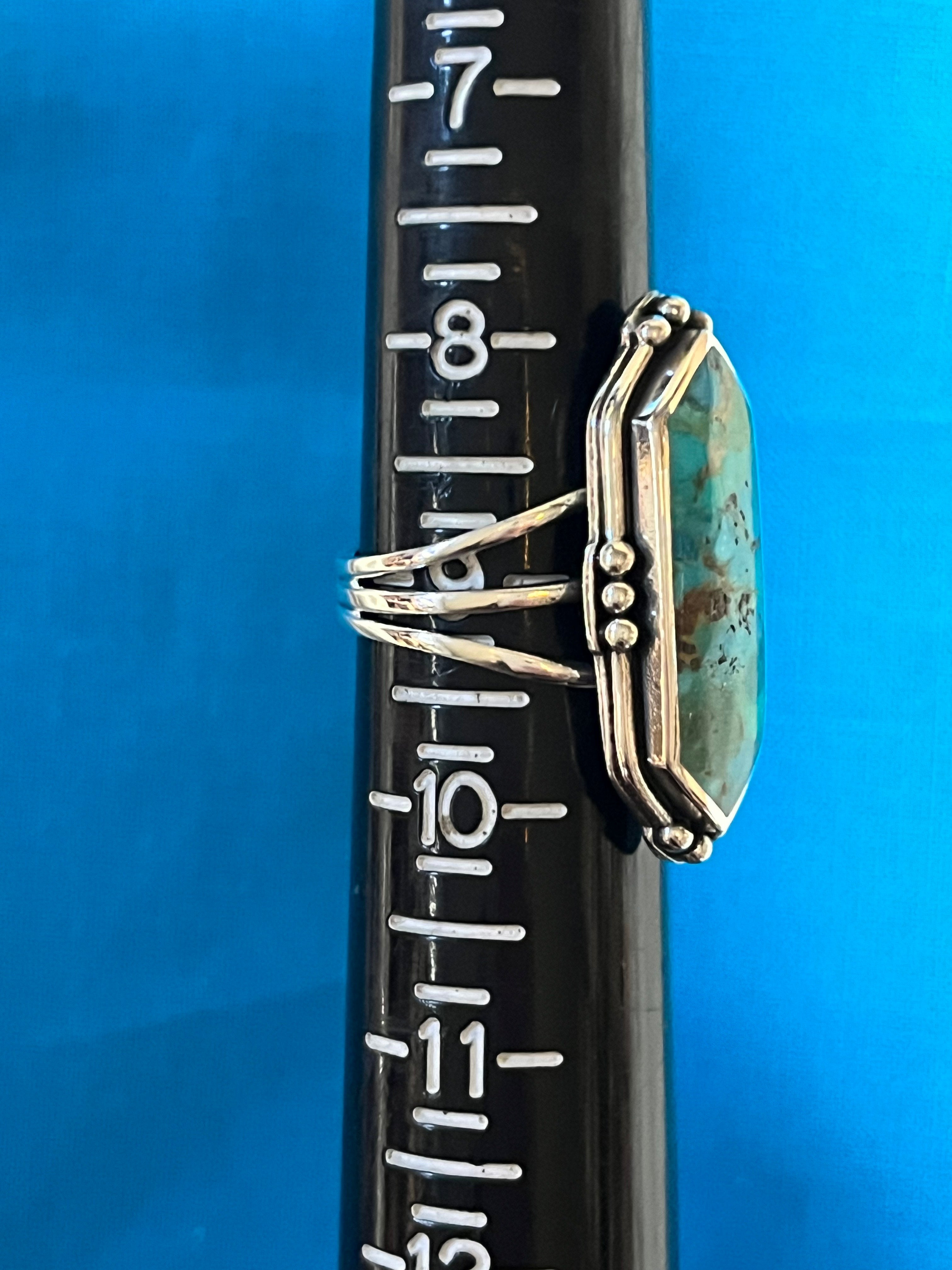 Silver ring with a turquoise stone on a black rod with measurements against a blue background